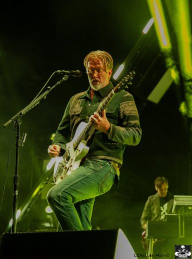 Musician performing on stage with a guitar, vibrant green lighting in the background.
