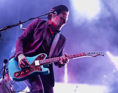 Musician in a suit playing a blue electric guitar on stage with vibrant lighting.