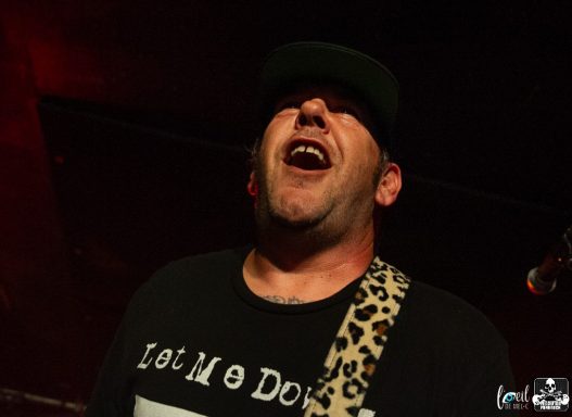 Man singing passionately on stage, wearing a black shirt and a leopard print guitar strap.