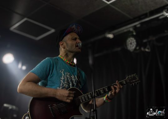Straightline - L'Anti-Bar & Spectacles Musician performing on stage, singing into a microphone while playing guitar.