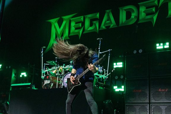 A guitarist performing energetically on stage with a drum set in the background.