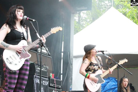 Two female musicians perform on stage with guitars, one wearing a black tank top and plaid pants.