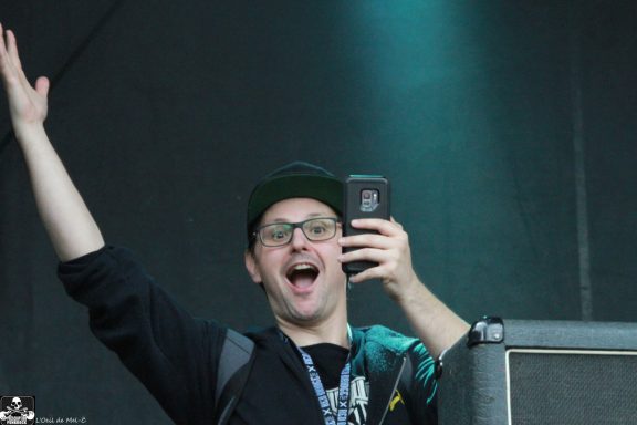 Person with glasses and a cap, smiling and waving while holding a phone in one hand.