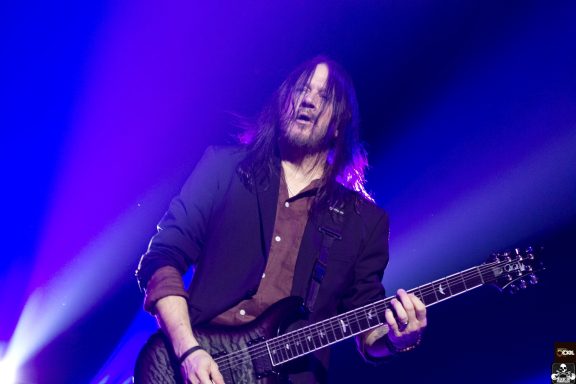 Musician performing on stage, holding a guitar with colorful lighting background.