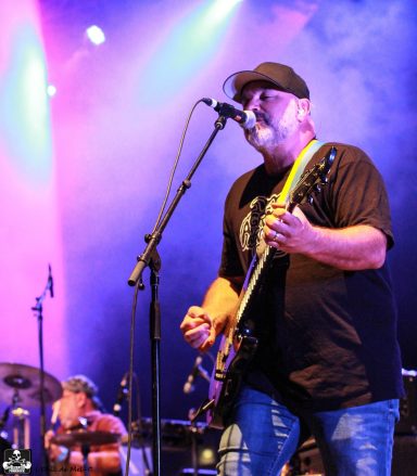 Musician performing on stage with guitar, drummer visible in the background.