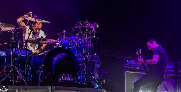 Drummer performing with a guitarist on stage, illuminated by colorful lights.