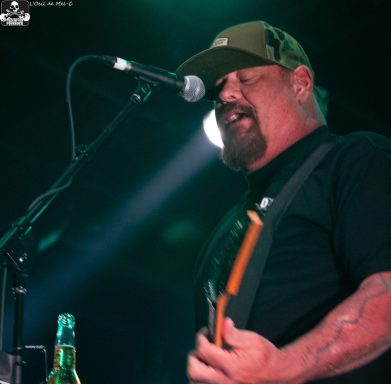 Musician performing on stage with a guitar and microphone, wearing a cap and black shirt.