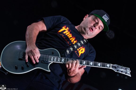 Musician playing an electric guitar on stage, wearing a black cap and a Thrash shirt.