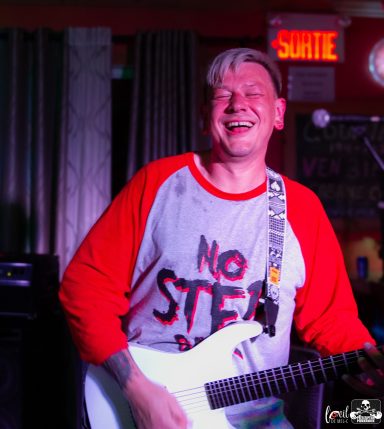 Smiling musician in a red and white shirt playing an electric guitar on stage.