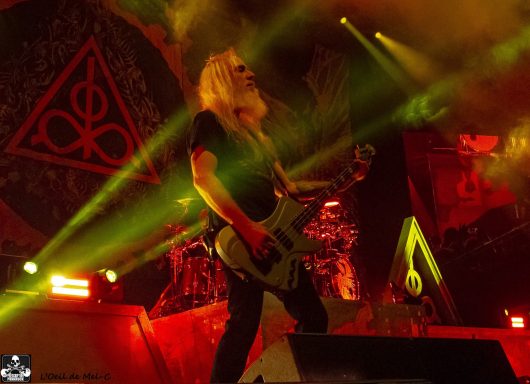 Musician playing bass guitar on stage with dramatic lighting and a backdrop design.