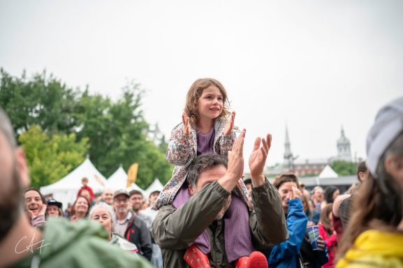 A young girl claps while sitting on a man's shoulders at an outdoor event.