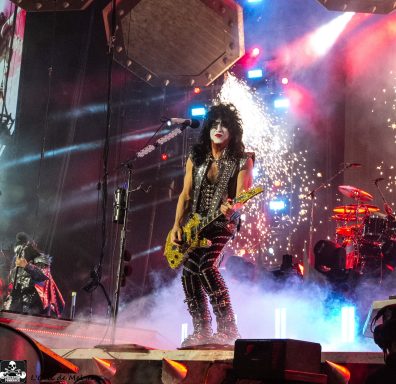 A guitarist performs on stage with pyrotechnics and colorful lights in the background.