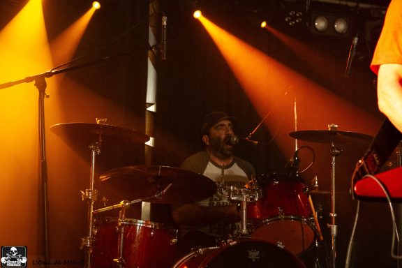 Drummer performing on stage with dramatic lighting in the background.