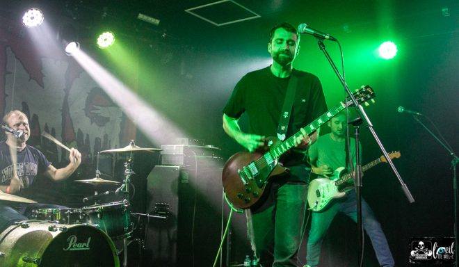 Band performing live on stage with green lighting and musical instruments.
