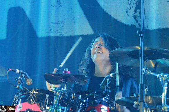 A drummer smiling while playing, with colorful lighting in the background.