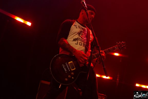 A musician performing on stage, playing an electric guitar with red stage lighting.