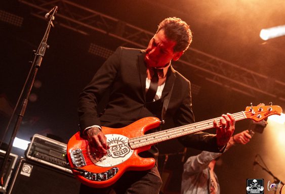 Musician in a suit playing an orange bass guitar on stage with dramatic lighting.