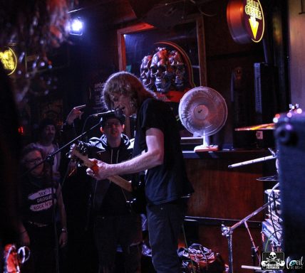 A musician performing on stage with a crowd and a fan in the background.