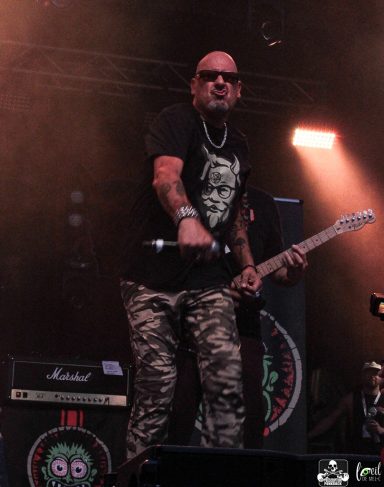 Musician performing on stage, wearing sunglasses and a graphic t-shirt, with a guitar.