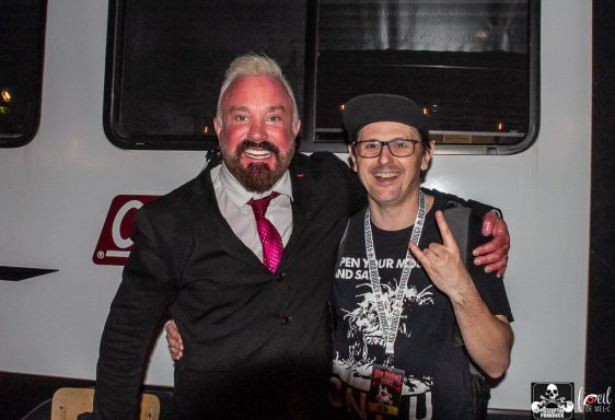 Two men pose together, one in a suit with a pink tie, the other in casual attire.