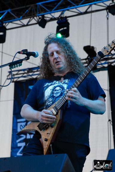 Musician playing electric guitar on stage, wearing a black shirt with graphic design.