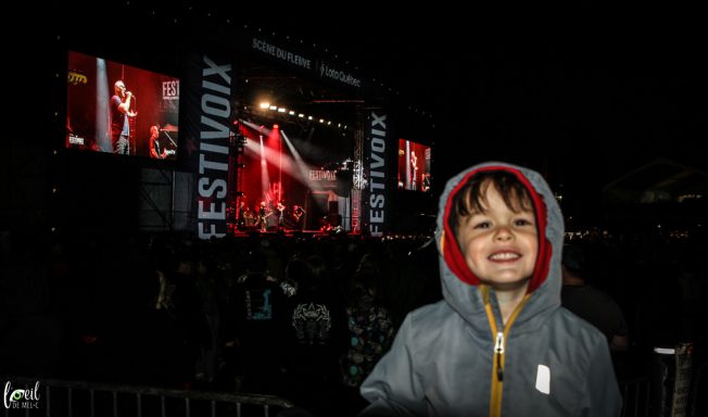 Smiling child in a hooded jacket at a nighttime music festival with a large stage backdrop.