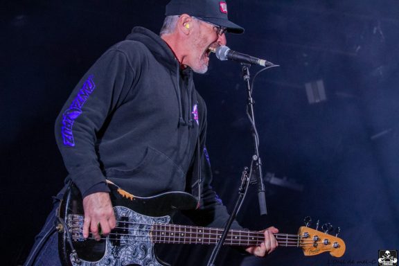 Musician in a black hoodie singing passionately while playing an electric bass guitar.