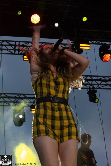 Singer in a yellow plaid jumpsuit performs on stage with bright lights in the background.