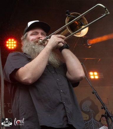 Man with a long beard playing a trombone on stage, with stage lights in the background.