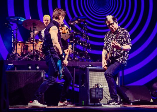 Two musicians perform on stage with drums and amplifiers in a colorful, swirling backdrop.