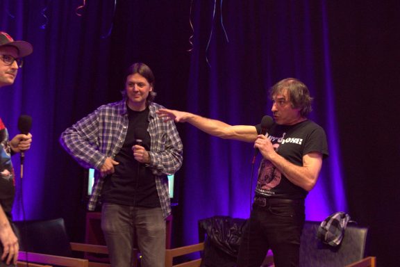 Two people on stage, one adjusting his shirt while the other gestures toward him.