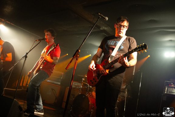 Two musicians perform on stage, one singing and the other playing guitar, with dramatic lighting.
