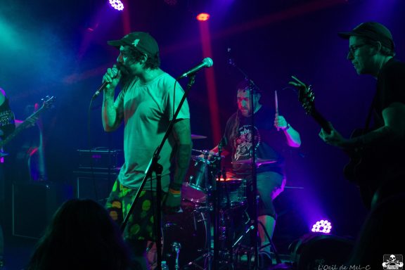 Band performing on stage with colorful lights, featuring a singer, drummer, and guitarist.