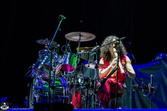 Musician playing drums on stage, wearing a red outfit with colorful lighting.