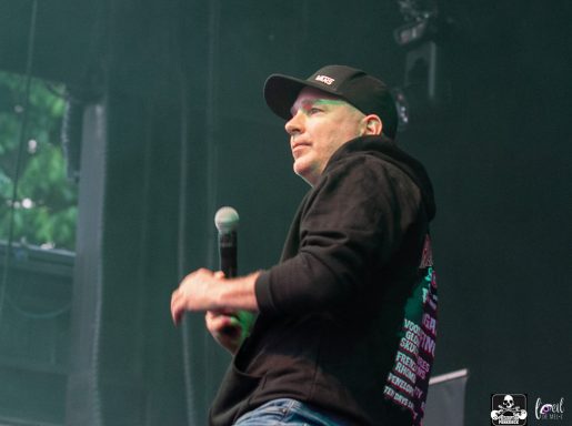 A man wearing a black cap and hoodie speaks into a microphone on stage.