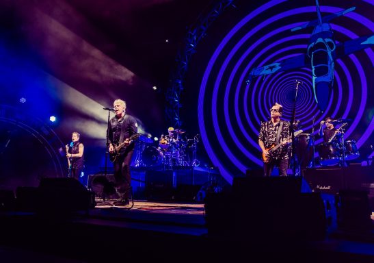 Band performing on stage with colorful lighting and circular visual effects.