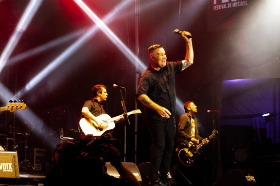 A performer sings on stage, with band members and bright stage lights in the background.