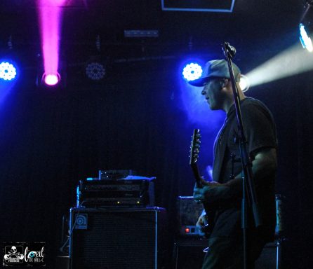 Guitarist performing on stage with dramatic lighting in the background.