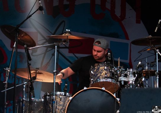 Drummer performing on stage with a colorful background and dramatic lighting.