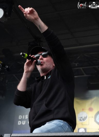 A performer wearing sunglasses and a hat gestures while singing into a microphone.
