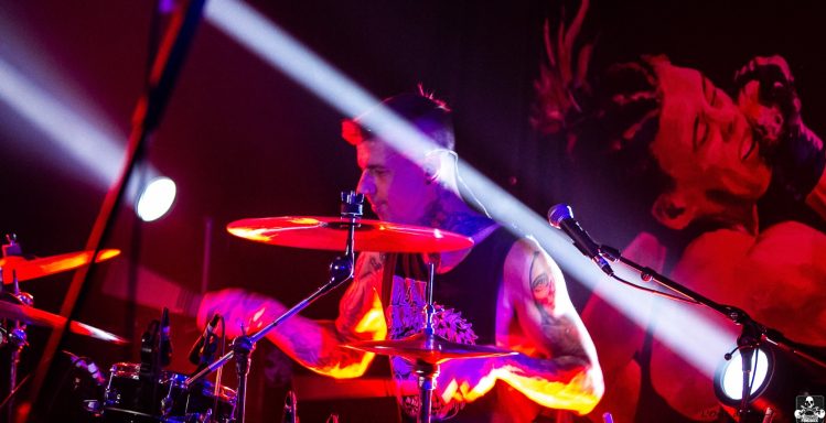 Drummer performing on stage with vibrant lighting and a mural in the background.