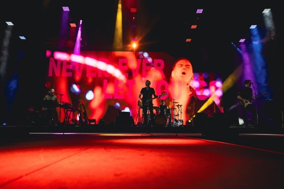 A band performing on stage with vibrant lighting and a large screen display in the background.