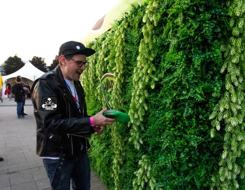 Man in a leather jacket waters a lush green wall of plants.