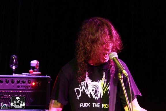 Musician performing on stage with long hair and a microphone, wearing a graphic t-shirt.