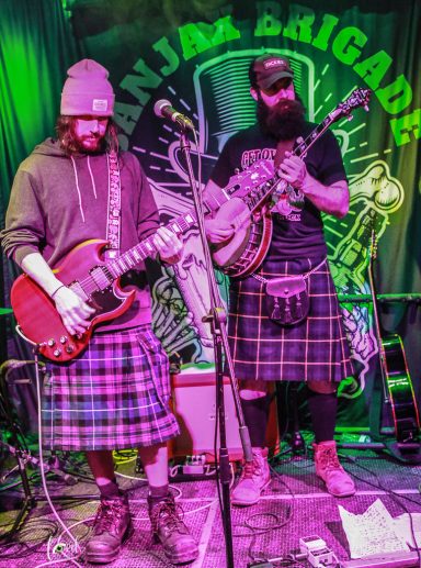 Two musicians in kilts perform on stage, one playing guitar and the other a banjo.
