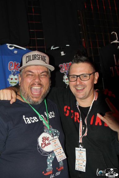 Two smiling men pose together, one wearing a cap and the other with glasses.