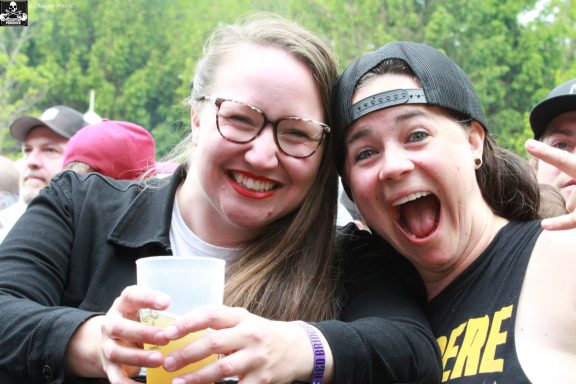 Two women smiling joyfully at a festival, holding drinks and posing playfully.