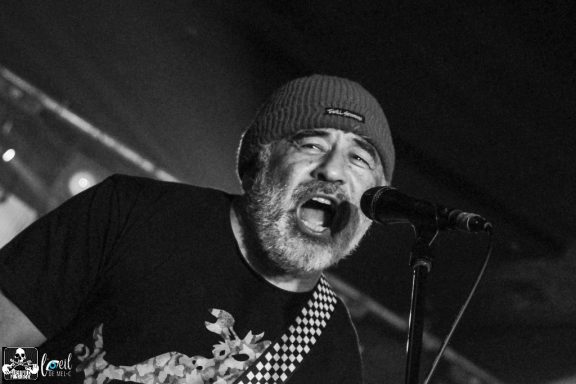 Man singing passionately into a microphone, wearing a beanie and guitar strap.