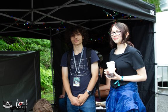 Two people stand under a tent; one is holding a cup and smiling.