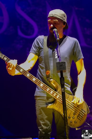 Musician performing on stage, holding a bass guitar and singing into a microphone.